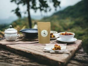 Dieses Bild zeigt unseren AY Natural Doypack gefüllt mit dem aromatischen Tee aus Vietnam auf einem charmanten Holztisch platziert Neben dem Doypack liegen Teller und Tassen gefüllt mit losem Tee was die Vielfalt und Qualität der Teeauswahl unterstreicht Im Hintergrund erstrecken sich grüne Gebirge die die natürliche Schönheit und Ursprünglichkeit Vietnams einfangen