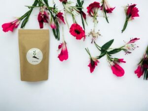 Dieses Bild zeigt unseren AY Natural Doypack gefüllt mit einer Auswahl an blumigen Tee Geschmacksnoten auf einem klaren weißen Hintergrund der von pinken Blumen verziert wird Die blumigen Aromen und Düfte der Tees werden durch die umgebenden Blumen betont und versprechen ein erfrischendes und duftendes Genusserlebnis