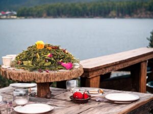Auf einem runden Holzbrett liegt der AY Natural loser Tee Die hellgrünen gerollten Blätter sind mit getrockneten Früchten gemischt Im Hintergrund ist ein See zu sehen davor liegt Besteck auf dem Tisch Es handelt sich um den Grüntee aus dem Himalaya