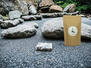 Hier präsentiert sich unser AY Natural Doypack auf einem Steinboden gefüllt mit einer Auswahl an Tee aus Japan Im Hintergrund erstrecken sich viele Steine und Felsen die die landschaftliche Schönheit Japans einfangen Tauche ein in die Welt der japanischen Teesorten und erlebe die Harmonie und Ruhe die sie bieten