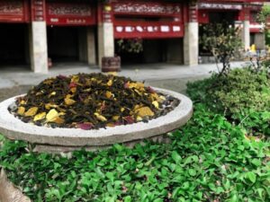Der AY Natural Pu Erh Tee gemischt mit gelben Blättern ruht hier auf einem Stein zwischen üppigen Pflanzen während im Hintergrund die geschäftige Stadt zu sehen ist Tauche ein in die entspannende Wirkung dieses chinesischen Tees und genieße eine Auszeit vom Trubel des Alltags