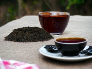 Hier liegt unser AY Natural Schwarztee aus Assam auf dem Tisch präsentiert in gerollten dunkelgrünen Blättern Neben dem Tee steht eine Tasse bereit für einen aromatischen Schluck Im Hintergrund erstrahlt die grüne Natur die die Frische und Reinheit dieses besonderen Schwarztees unterstreicht