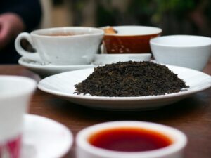Hier liegt unser AY Natural Schwarzer Tee auf einem Tisch serviert in einem dunkelgrünen Teller Im Hintergrund sind Tee Tassen zu sehen die auf den Genuss dieses chinesischen Schwarztees warten Tauche ein in die Welt der Grüntöne und erlebe den aromatischen Geschmack dieses besonderen Tees aus China