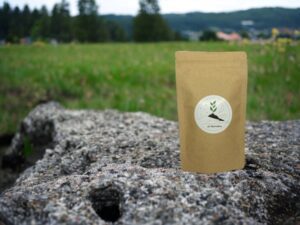Erlebe die Ruhe und Harmonie der Natur mit AY Natural Unser Doypack präsentiert sich stolz auf einem Steinfelsen gefüllt mit unserem feinen losen Tee Im Hintergrund erstreckt sich eine idyllische grüne Wiese gesäumt von majestätischen Bäumen