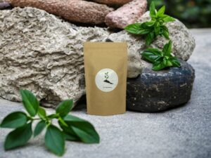 Dieses Bild zeigt unseren AY Natural Doypack gefüllt mit der belebenden Tulsi Teemischung vor einem imposanten Steinfelsen Die natürliche Schönheit der Umgebung wird durch die Anwesenheit von grünen Pflanzen verstärkt die die Frische und Reinheit unserer Tees unterstreichen Die Tulsi Teemischung vereint die belebenden Eigenschaften von Tulsi mit einer harmonischen Mischung aus anderen Kräutern und Gewürzen