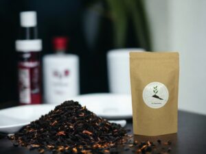 Schwarzer loser Tee liegt verstreut auf dem Tisch Daneben der brauner Doypack mit dem AY Natural Logo Im Hintergrund stehen Objekte Der Hintergrund ist verschwommen